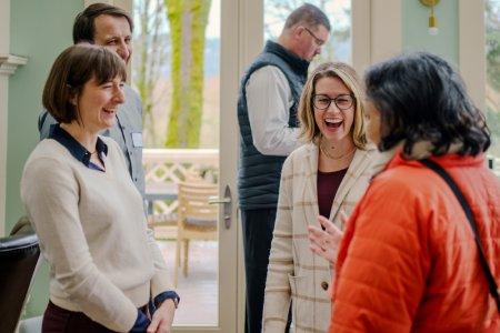 A group of 4 smiling and lauging coworkers having an engaging conversation indoors. There is a man in the background focused on something else.